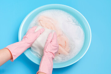 Female hands in gloves washes clothes with powder in basin on blue background close-up top view.