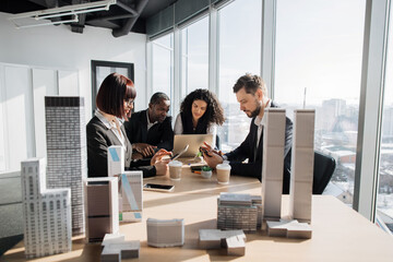 Business team of four good-looking multiethnic corporate workers discussing financial report using laptop and gadgets working on building complex prototype project of residential or business district