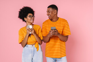 Canvas Print - African man and woman using mobile phones and looking at each other while standing isolated over pink wall