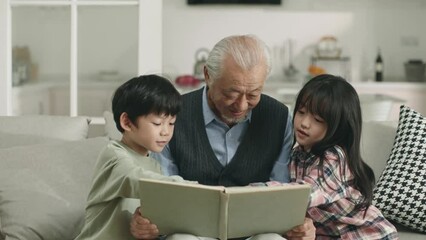 Wall Mural - asian grandpa and two grandchildren sitting on couch at home reading a book together