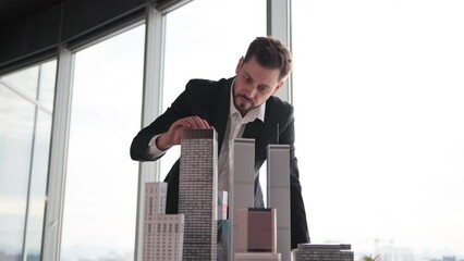 Wall Mural - Confident businessman real estate agents in black stylish formal suit standing while touching 3d model skyscraper of city architecture design at office with panoramic city view.