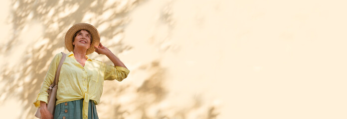 happy senior woman in sun hat walks  on summer city against the background of a yellow wall with shadows from trees. banner