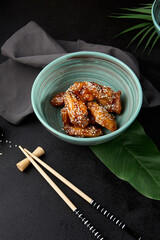 Poster - Roasted chicken wings in teriyaki sauce with sesame on ceramic bowl. Buffalo wings in asian style on black concrete background. Aesthetic composition with fried chicken wings, chopsticks and leaf.