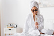 © LIGHTFIELD STUDIOS - happy african american woman in terry bathrobe and trendy sunglasses sitting on bed with glass of champagne.