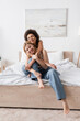 happy african american woman embracing blonde barefoot friend in jeans sitting on bed at home.