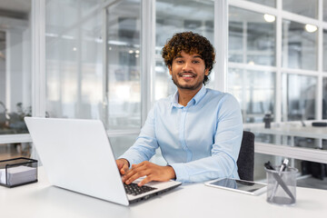 Portrait of young successful contemporary businessman inside office, man at workplace smiling and looking at camera, young hispanic businessman sitting at desk using laptop.