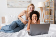 © LIGHTFIELD STUDIOS - excited multiethnic friends in lingerie and jeans watching film on laptop in bedroom.