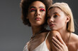 © LIGHTFIELD STUDIOS - blonde and brunette interracial women with bright visage and silver necklaces posing isolated on grey.