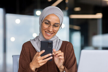 Wall Mural - Close up of muslim successful businesswoman in hijab using phone inside modern office, workplace woman typing message and typing on smartphone screen smiling.