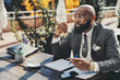 © skyNext - A black balding corporate man with a beard sitting in an outdoor cafe, dressed in a fancy suit, polka-dot tie, and a white vest, reviews his notes with a pen, and while taking a sip of his coffee