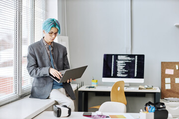 Portrait of young man with colored blue hair using laptop while sitting by window in office, copy space