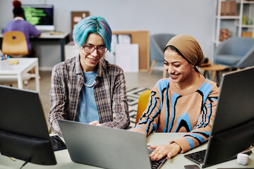 Portrait of two young software developers using computers in office and smiling happily enjoying work