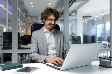 Cheerful and successful businessman inside office working with laptop, bearded man smiling and typing on laptop keyboard, programmer coding software in modern room.