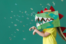 Little Boy In Cardboard Dinosaur Costume And Soap Bubbles On Green Background