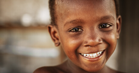 Wall Mural - Close Up Portrait of a Shy Authentic African Boy Smiling and Laughing at the Camera with a Blurred Background. Black Male Kid Representing Future, Hope, and Acceptance. Documentary Footage