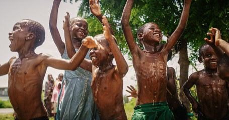 Wall Mural - Close Up on a Group of Happy and Innocent Black Children Playing and Enjoying the Blessing of Rain Water After Long Drought. Authentic African Kids Jumping and Laughing when Water Gets Poured on Them.