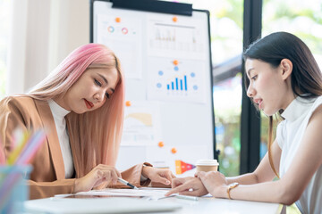 Wall Mural - Two pretty young Asian businesswoman sitting at desk with laptop doing paperwork together discussing project financial report. Corporate business collaboration concept.