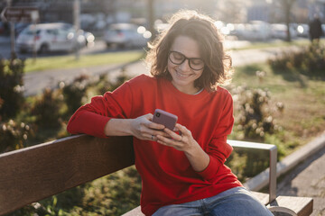 Young curly beautiful woman hipster in glasses smiling and texting using smartphone sitting on bench in city