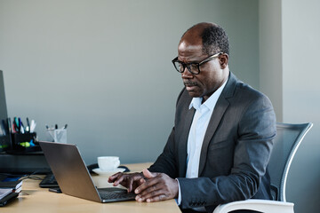 Poster - Serious mature African American businessman in formalwear and eyeglasses using laptop while sitting by desk in his office