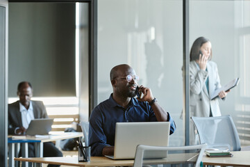 Fototapete - Young confident businessman with smartphone by ear talking to client while sitting by workplace in front of laptop against his coworkers