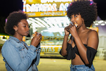 Two Young Black Girls With Afro Hair In Jeans Clothing Eating Hamburger Near Food Truck At Night