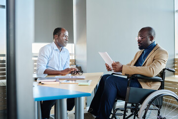 Poster - Young male economist with disability making report to mature African American boss sitting by his workplace in front of subordinate