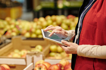 Wall Mural - Close-up of mature female administrator of supermarket in red uniform using tablet while looking through list of goods during revision