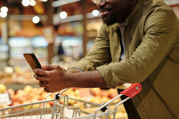 Side view close-up of young male consumer with smartphone over shopping cart watching online video about some goods