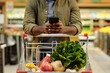 © pressmaster - Young African American male consumer pushing shopping cart with fresh vegetarian food products and scrolling in smartphone in supermarket
