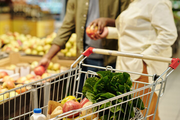 Wall Mural - Selective focus on shopping cart with fresh fruits and vegetables on background of young couple choosing apples in grocery store
