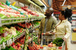 © pressmaster - Young African American couple of shoppers standing by large display with fresh vegetables and choosing tomatoes and other products