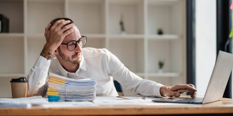 Wall Mural - Young unhappy man office worker feeling bored at work, looking at laptop with demotivated face expression while sitting at workplace in office, distracted male worker feeling tired of monotonous job