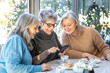 © Andrea Vumbaca Photo - Group of 3 smiling mature female friends in white hats are using smartphone while at cafe having breakfast . Three senior women laugh look at the phone together