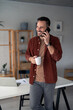 © Dorde - Cheerful modern man freelancer on cell phone conversation standing in front of table with laptop and paper document on it in home office room, looking away and smiling. Coffee break in home office.