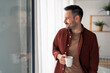 © Dorde - Satisfied smiling man standing indoors, holding white cup of coffee and looking away through window with white curtains. Happy man enjoying coffee at home.