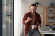 © Dorde - Handsome good-looking man, modern entrepreneur in stylish casual clothes cheerfully looking at mobile phone screen and holding white cup of coffee while sitting on the edge of the table in home office