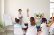 © Studio Romantic - Group of elementary school students raising hands to answer question in classroom. Cute children sitting on chairs with their backs to camera. Cheerful female teacher conducting lesson at school