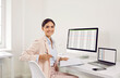 © Studio Romantic - Portrait of a young happy business woman sitting at the desk on workplace, looking at the camera and smiling. Cheerful accountant working on a laptop with tables and charts at office or at home.