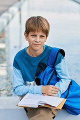 Wall Mural - Cute secondary school learner with open book looking at camera while sitting in urban environment and preparing for lesson of literature