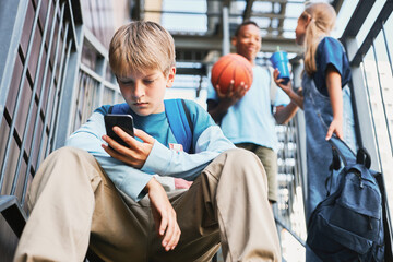 Wall Mural - Adolescent schoolboy with backpack scrolling in smartphone while sitting on playground facilities against two intercultural friends