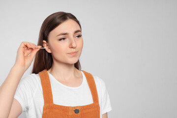 Poster - Young woman cleaning ear with cotton swab on light grey background. Space for text