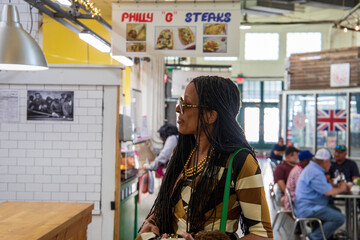 An African American woman with long sisterlocks wearing a brown with and black dress, sunglasses, and a green purse buying food in a food hall at the Municipal Market in Atlanta Georgia USA