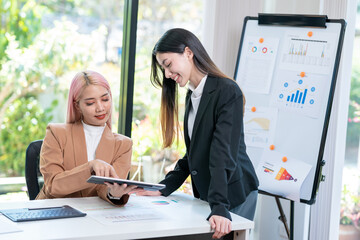 Wall Mural - Two young Asian business women meet to analyze the financial chart at the office to discuss the financial situation at the company. A partner sits at a desk with modern documents and equipment.