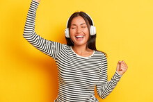 People, Music, Emotions Concept. Horizontal Shot Of Pretty Asian Young Woman Carried Away With Music Dances Joyfully In White Headphones With Closed Eyes Smiles Cheerfully Wears Striped Jumper