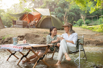 Wall Mural - Pleased happy mother and daughter reading a book and using laptop while relaxing on the deck chairs in the river, sit near a camp and tent, drink coffee in a pine forest. Camping, recreation, hiking.