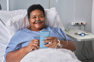 Portrait of smiling senior woman using phone while laying on bed in hospital room