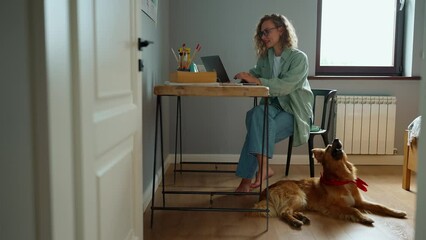 Wall Mural - Lovely curly haired woman working on laptop and stroking her dog lying on the floor at home