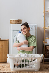 Wall Mural - Positive woman holding washing liquid and looking at camera in laundry room.