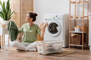 Wall Mural - Side view of cheerful brunette woman holding clothes near basket in laundry room.