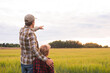 © Acronym - Farmer and his son in front of a sunset agricultural landscape. Man and a boy in a countryside field. Fatherhood, country life, farming and country lifestyle concept.